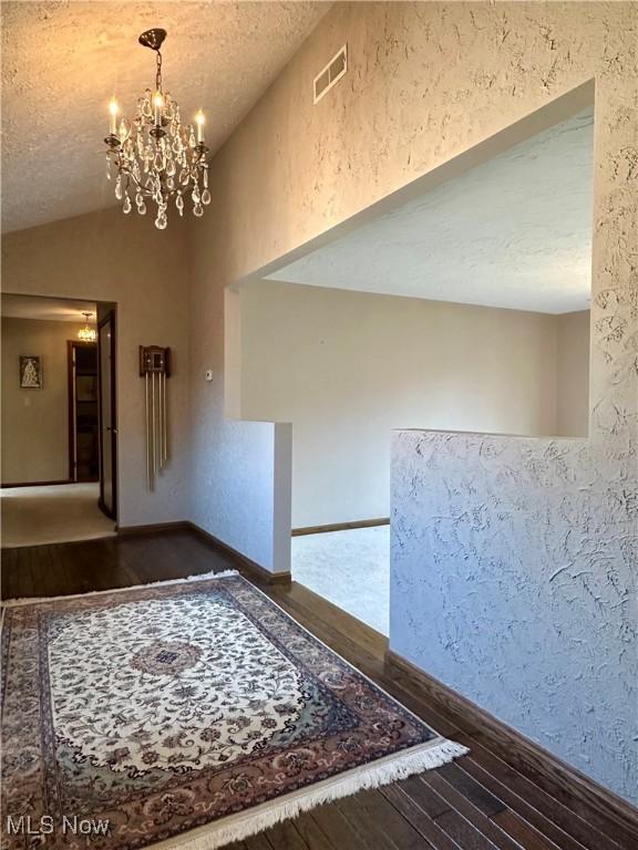 empty room with visible vents, wood-type flooring, a chandelier, vaulted ceiling, and a textured wall