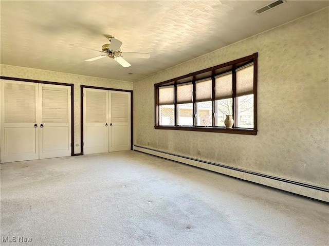 unfurnished bedroom with a ceiling fan, visible vents, baseboard heating, light colored carpet, and two closets