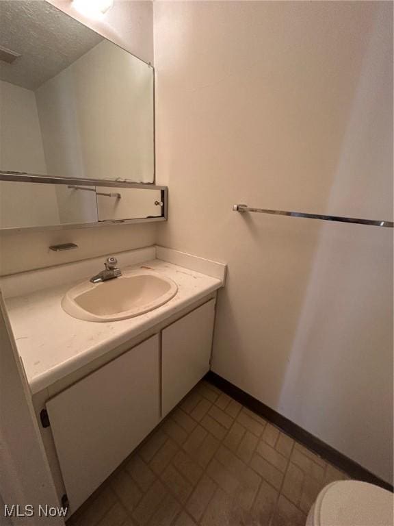 half bath with baseboards, toilet, vanity, brick floor, and a textured ceiling