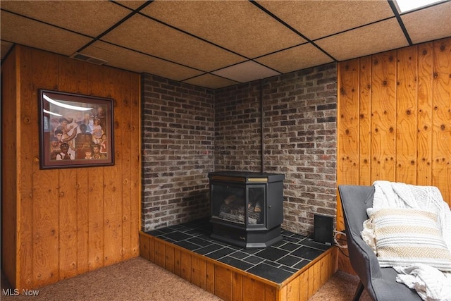 room details with visible vents, a drop ceiling, wooden walls, carpet, and a wood stove