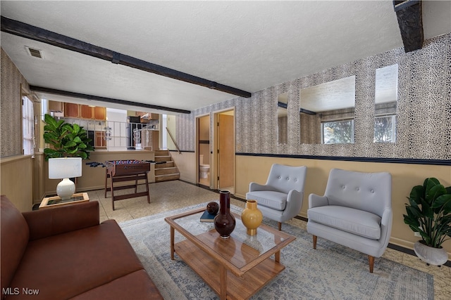 living area with beamed ceiling, visible vents, a textured ceiling, wainscoting, and wallpapered walls