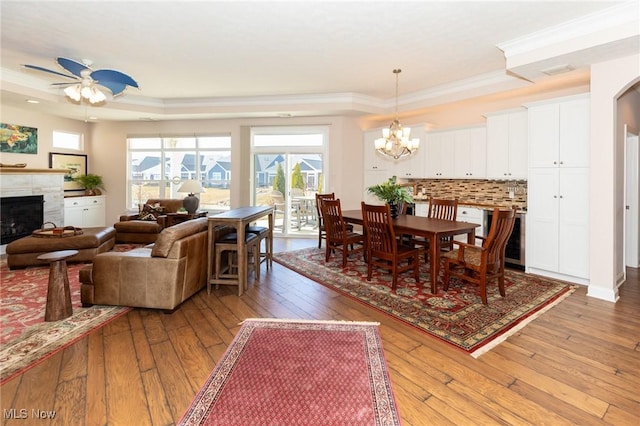 dining room with ceiling fan with notable chandelier, a tray ceiling, light wood-style floors, crown molding, and a premium fireplace