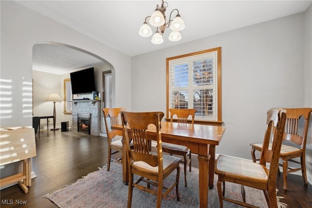 dining room with wood finished floors, arched walkways, a stone fireplace, baseboards, and a chandelier