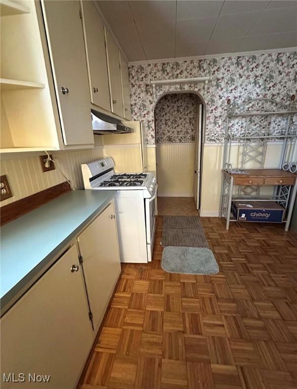 kitchen with wallpapered walls, under cabinet range hood, white range with gas stovetop, wainscoting, and white cabinetry