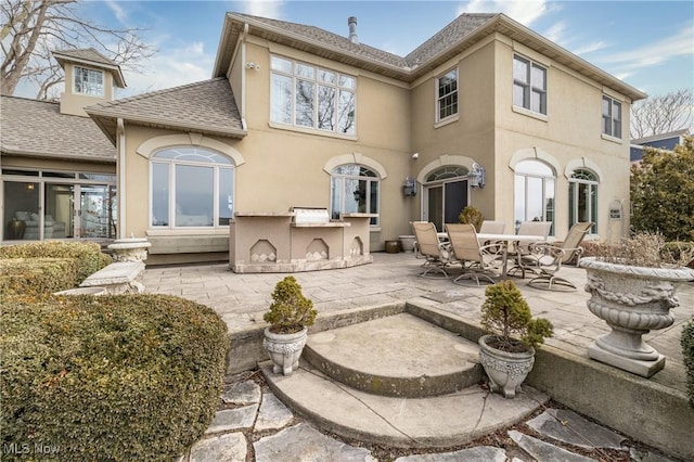 back of property with a shingled roof, a patio, an outdoor kitchen, and stucco siding