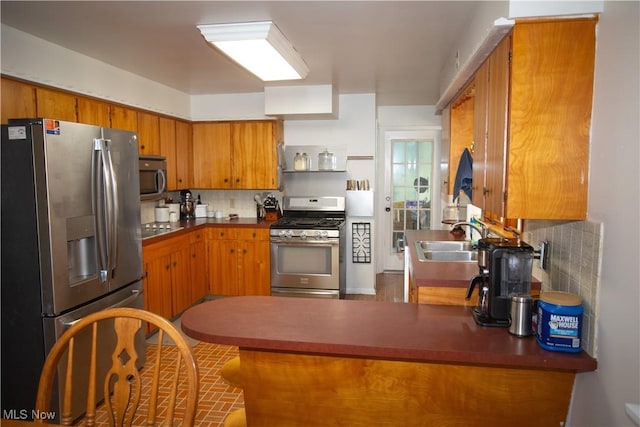 kitchen featuring brown cabinetry, a peninsula, a sink, decorative backsplash, and appliances with stainless steel finishes