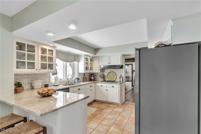 kitchen featuring a sink, light stone counters, under cabinet range hood, freestanding refrigerator, and a peninsula