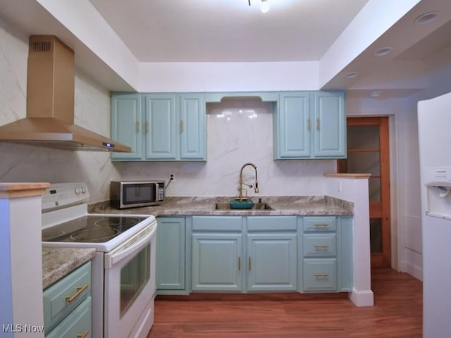 kitchen with a sink, blue cabinetry, dark wood-style floors, white appliances, and wall chimney range hood