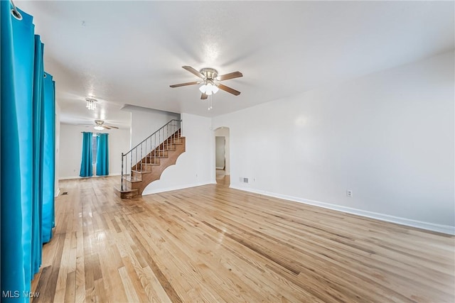 unfurnished living room with ceiling fan, baseboards, stairway, light wood-style floors, and arched walkways