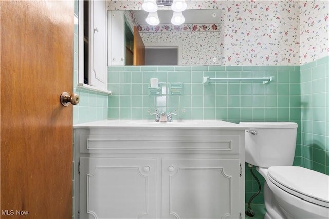 half bath featuring wallpapered walls, toilet, vanity, and a wainscoted wall