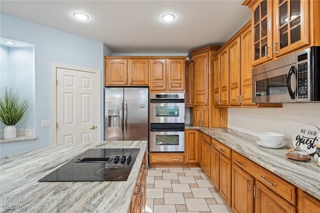 kitchen featuring brown cabinetry, light stone counters, glass insert cabinets, and appliances with stainless steel finishes