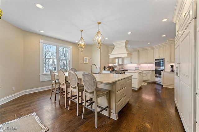 kitchen with custom exhaust hood, backsplash, an island with sink, and dark wood-type flooring