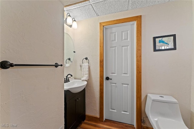 half bathroom featuring toilet, wood finished floors, a paneled ceiling, vanity, and a textured wall