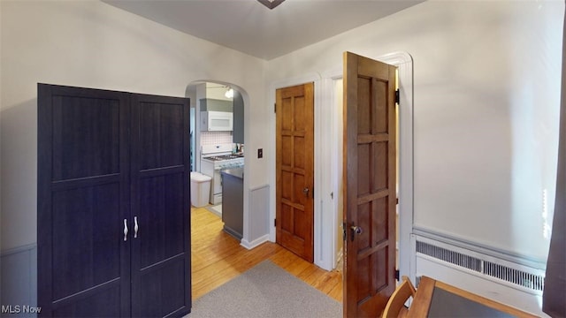 hall with arched walkways, light wood-style flooring, and radiator heating unit