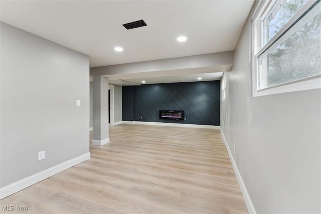 basement featuring an accent wall, baseboards, recessed lighting, light wood-style floors, and a glass covered fireplace