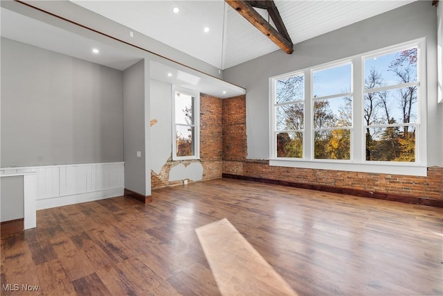 unfurnished living room with wood finished floors, brick wall, lofted ceiling with beams, recessed lighting, and wainscoting