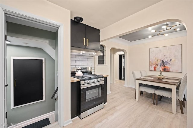 kitchen with stainless steel gas range oven, backsplash, under cabinet range hood, arched walkways, and dark cabinets