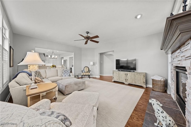 living area with a stone fireplace, ceiling fan with notable chandelier, baseboards, and wood finished floors