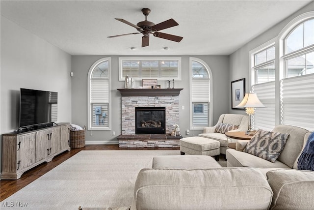 living room with baseboards, a stone fireplace, ceiling fan, and dark wood-style flooring