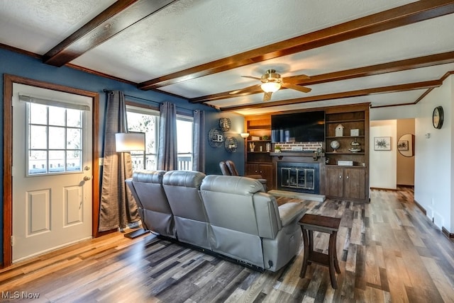 living area featuring a glass covered fireplace, beamed ceiling, wood finished floors, and ceiling fan