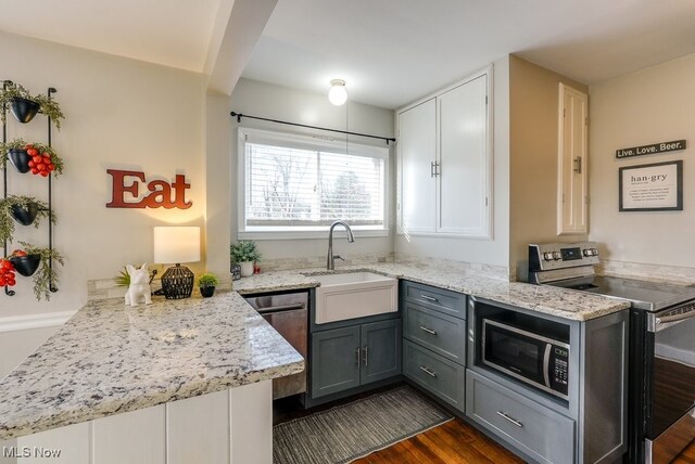 kitchen with gray cabinetry, light stone countertops, a peninsula, appliances with stainless steel finishes, and a sink