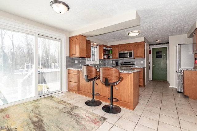 kitchen with light tile patterned floors, brown cabinets, a peninsula, stainless steel appliances, and tasteful backsplash