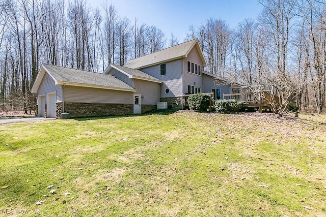 rear view of property with a shingled roof, a wooden deck, a garage, a yard, and stone siding