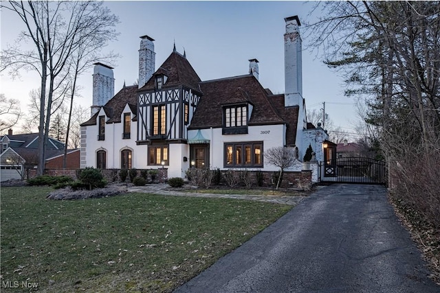 english style home featuring a front yard, a gate, stucco siding, a chimney, and aphalt driveway