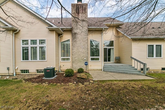 rear view of property with a lawn, cooling unit, roof with shingles, a chimney, and a patio area