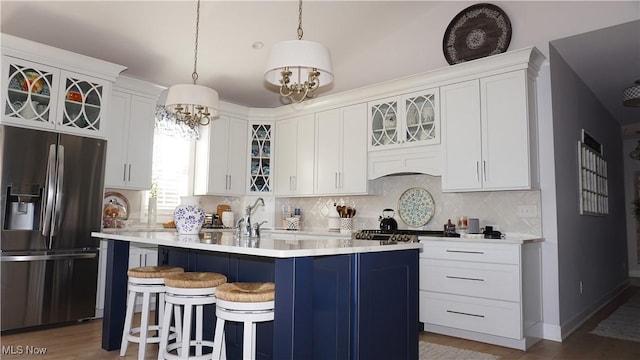 kitchen featuring a kitchen island with sink, backsplash, stainless steel fridge, light countertops, and glass insert cabinets