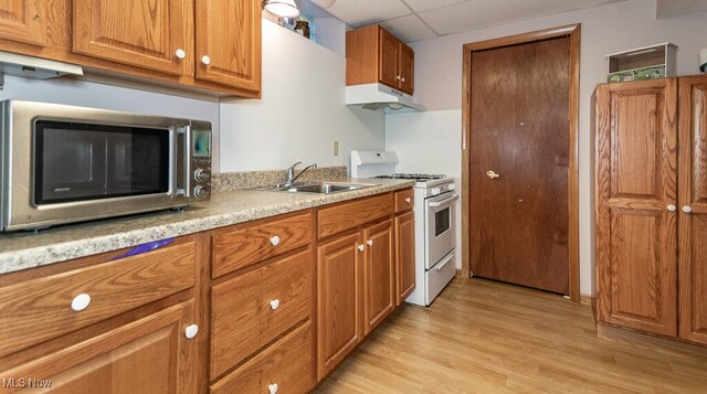 kitchen featuring stainless steel microwave, under cabinet range hood, white range with gas stovetop, brown cabinets, and a sink