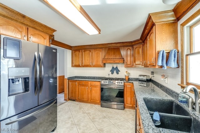 kitchen with premium range hood, decorative backsplash, appliances with stainless steel finishes, brown cabinetry, and a sink