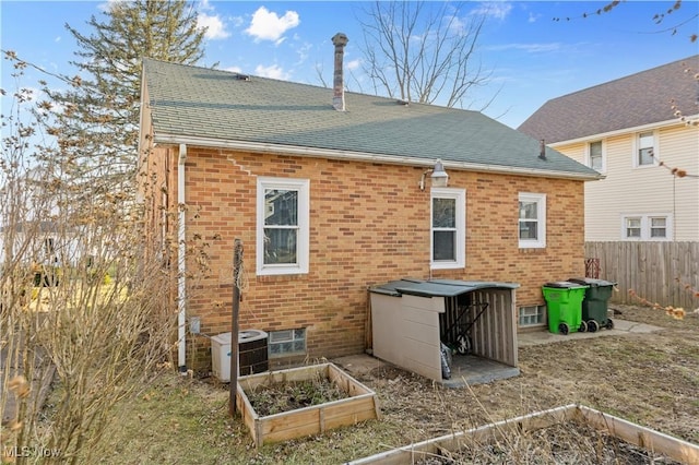 rear view of property featuring brick siding, a shingled roof, fence, central AC unit, and a vegetable garden