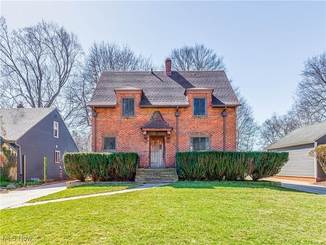 view of front facade with brick siding, a chimney, a front yard, and roof with shingles