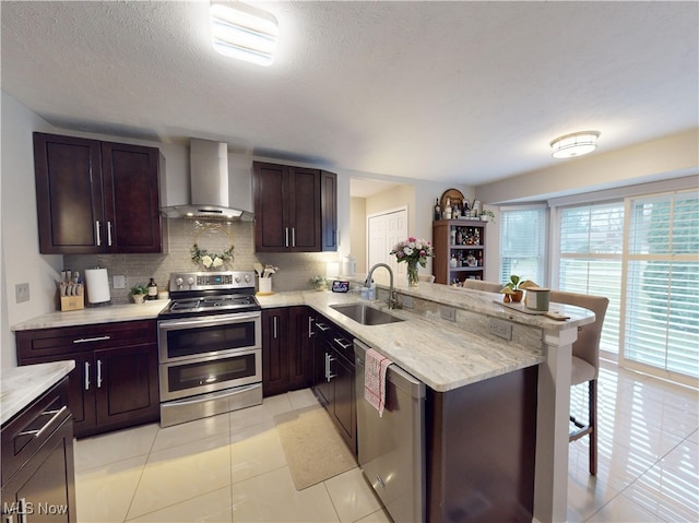 kitchen featuring a kitchen bar, a sink, stainless steel appliances, a peninsula, and wall chimney range hood