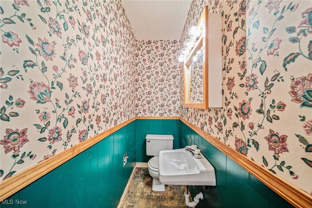 bathroom with wallpapered walls, toilet, a wainscoted wall, and a sink