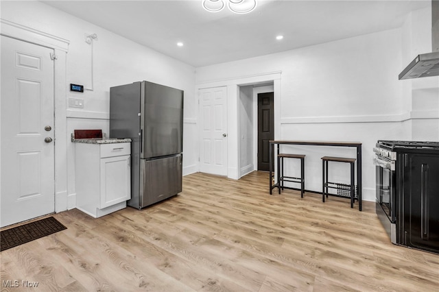 kitchen with light wood-style flooring, white cabinetry, stainless steel appliances, wall chimney range hood, and light stone countertops