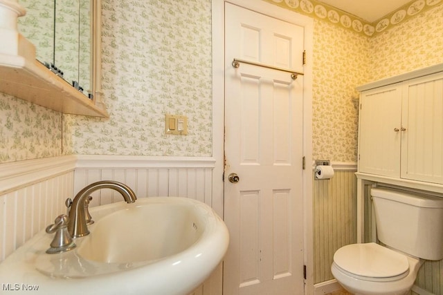 bathroom with wainscoting, wallpapered walls, toilet, and a sink