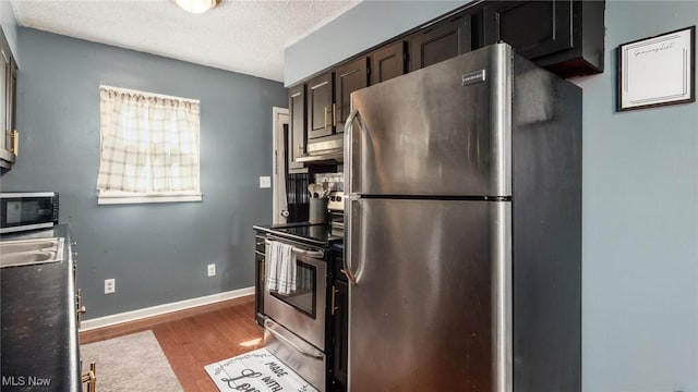 kitchen featuring wood finished floors, baseboards, dark brown cabinetry, appliances with stainless steel finishes, and a textured ceiling