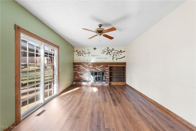 unfurnished living room with wood finished floors, a ceiling fan, visible vents, baseboards, and a brick fireplace