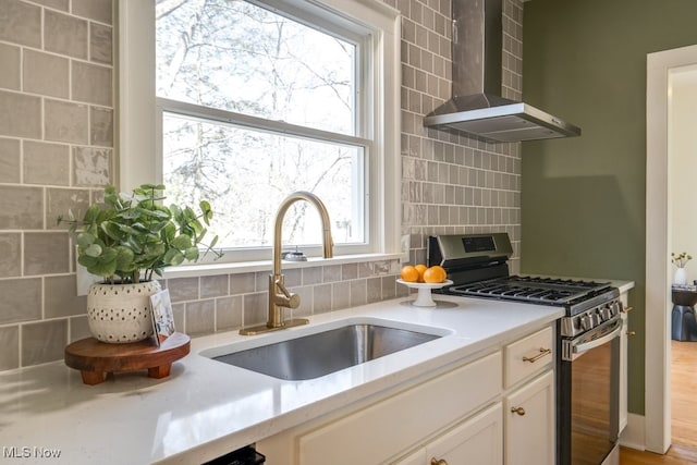 kitchen with stainless steel gas range, a sink, light countertops, wall chimney exhaust hood, and backsplash