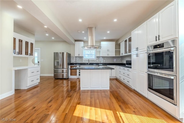 kitchen with island exhaust hood, stainless steel appliances, glass insert cabinets, light wood-type flooring, and backsplash