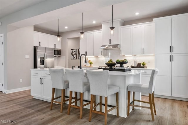 kitchen featuring light wood finished floors, under cabinet range hood, a center island with sink, appliances with stainless steel finishes, and a sink