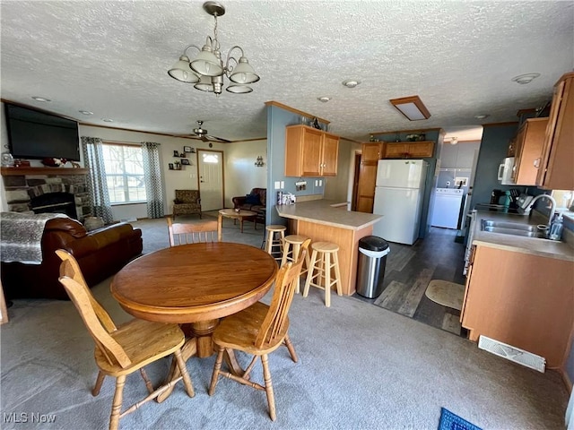 dining area featuring ceiling fan with notable chandelier, washer / clothes dryer, a textured ceiling, a stone fireplace, and dark colored carpet