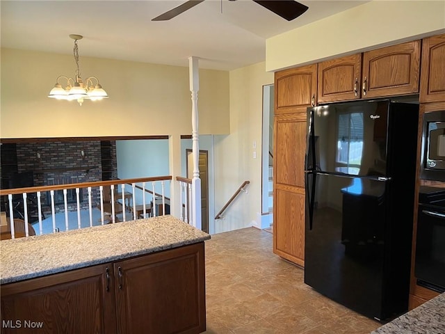 kitchen with light stone countertops, pendant lighting, ceiling fan with notable chandelier, brown cabinetry, and black appliances