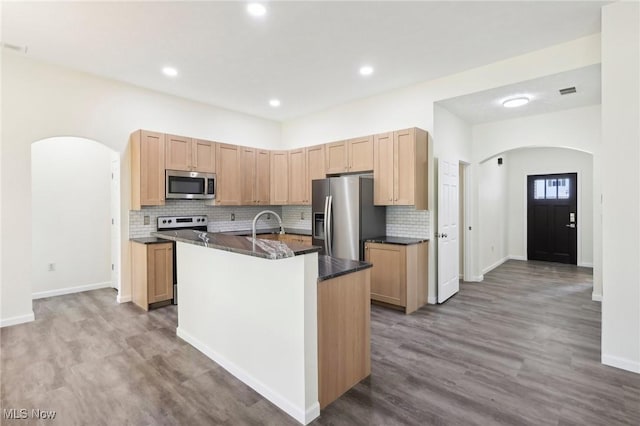 kitchen featuring wood finished floors, an island with sink, arched walkways, appliances with stainless steel finishes, and dark countertops