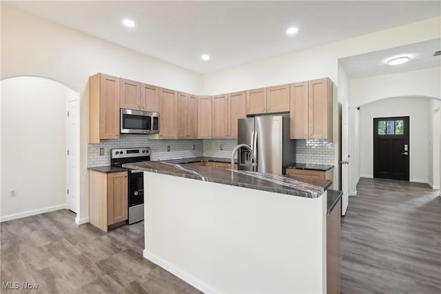 kitchen featuring a center island with sink, backsplash, dark countertops, arched walkways, and appliances with stainless steel finishes