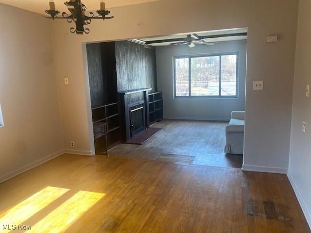 unfurnished living room with ceiling fan with notable chandelier, a fireplace with flush hearth, wood finished floors, and baseboards