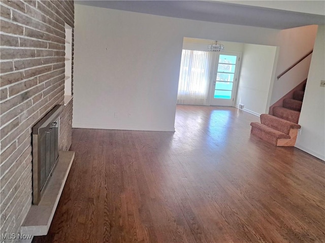 unfurnished living room with wood finished floors, brick wall, a brick fireplace, a chandelier, and stairs