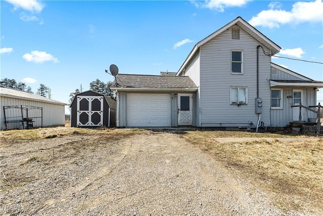 view of front facade with an outbuilding, an attached garage, a shingled roof, dirt driveway, and a storage shed